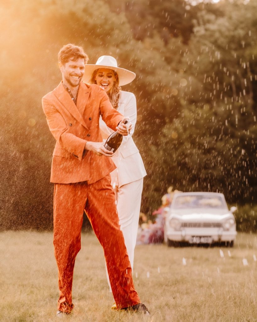 (C)HayleyKimHall A couple celebrates outdoors on a sunny day, captured brilliantly by a Sussex photographer. The man in a vibrant orange suit pops a champagne bottle, spraying bubbles as the woman in a white suit and hat laughs behind him. A vintage car is blurred in the background.