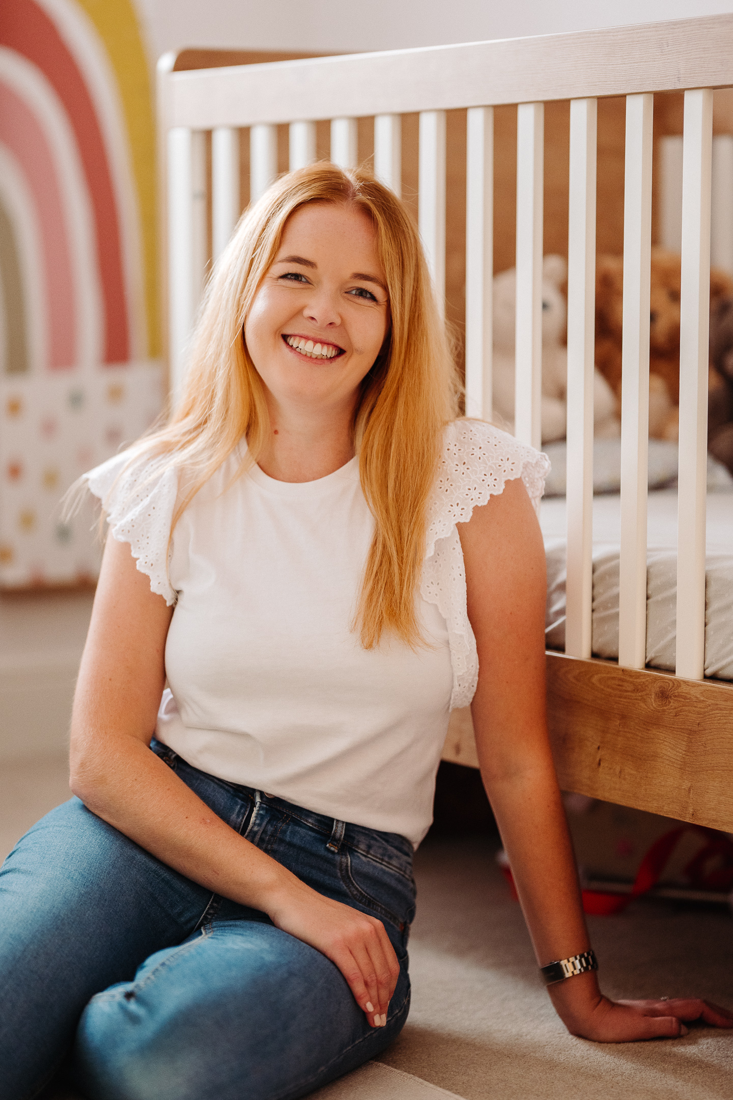 A smiling woman with long red hair sits on the floor beside a wooden crib.