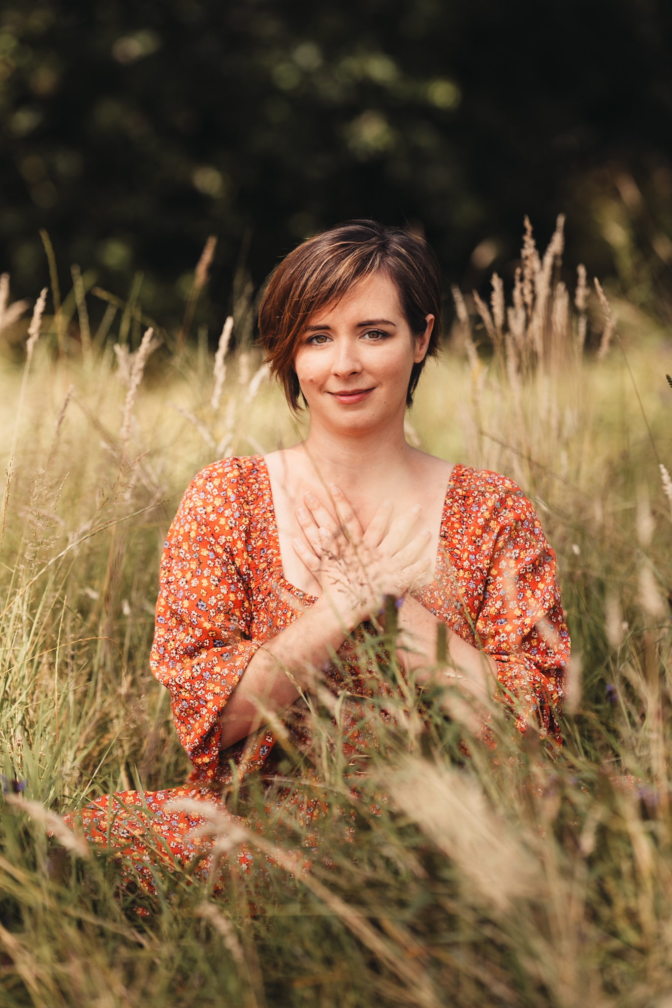 A woman sits in tall grass, wearing an orange floral dress.