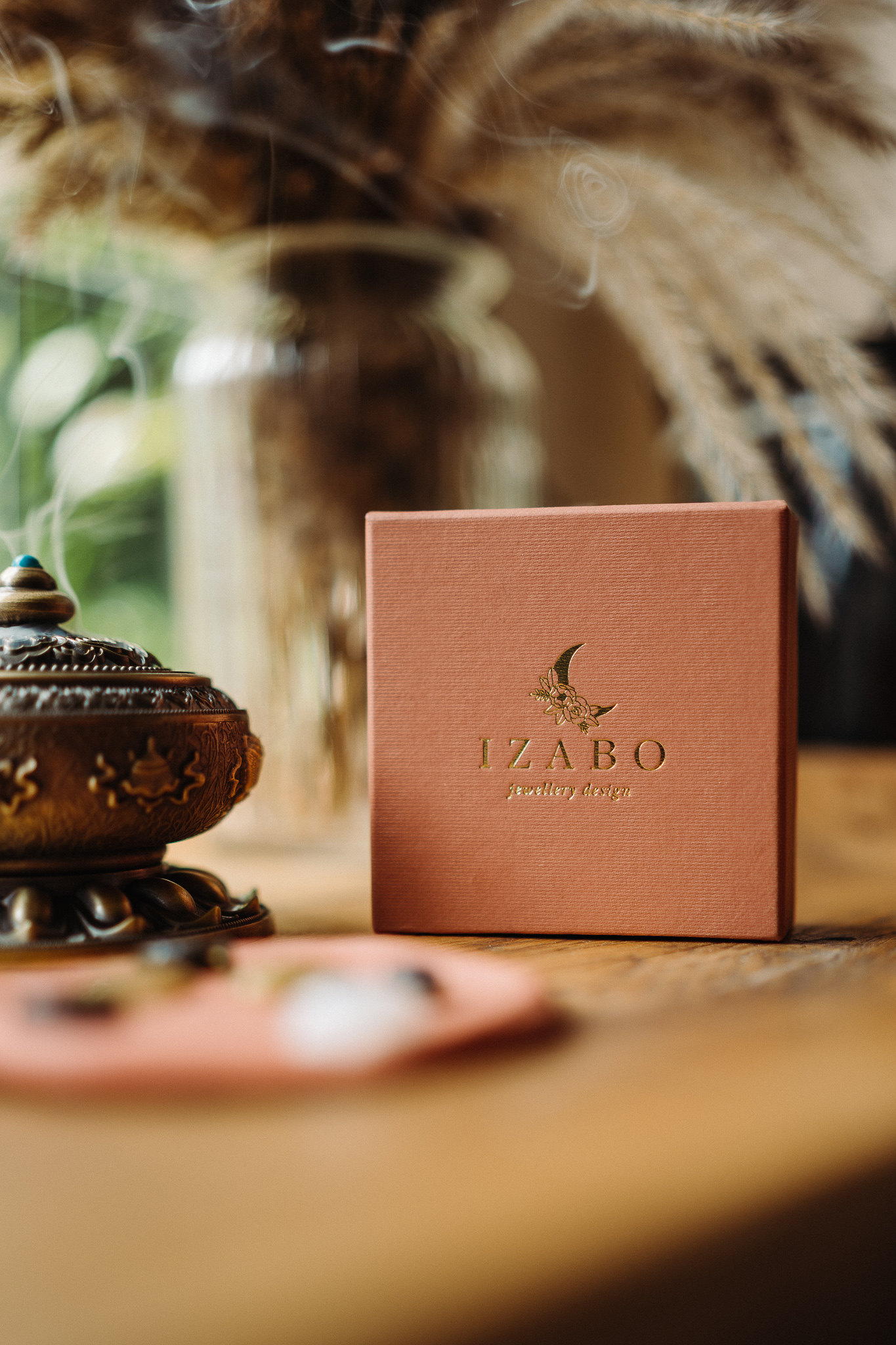 A peach-colored square box with gold embossed text and logo for Izabo Jewellery Design sits elegantly on a wooden table.