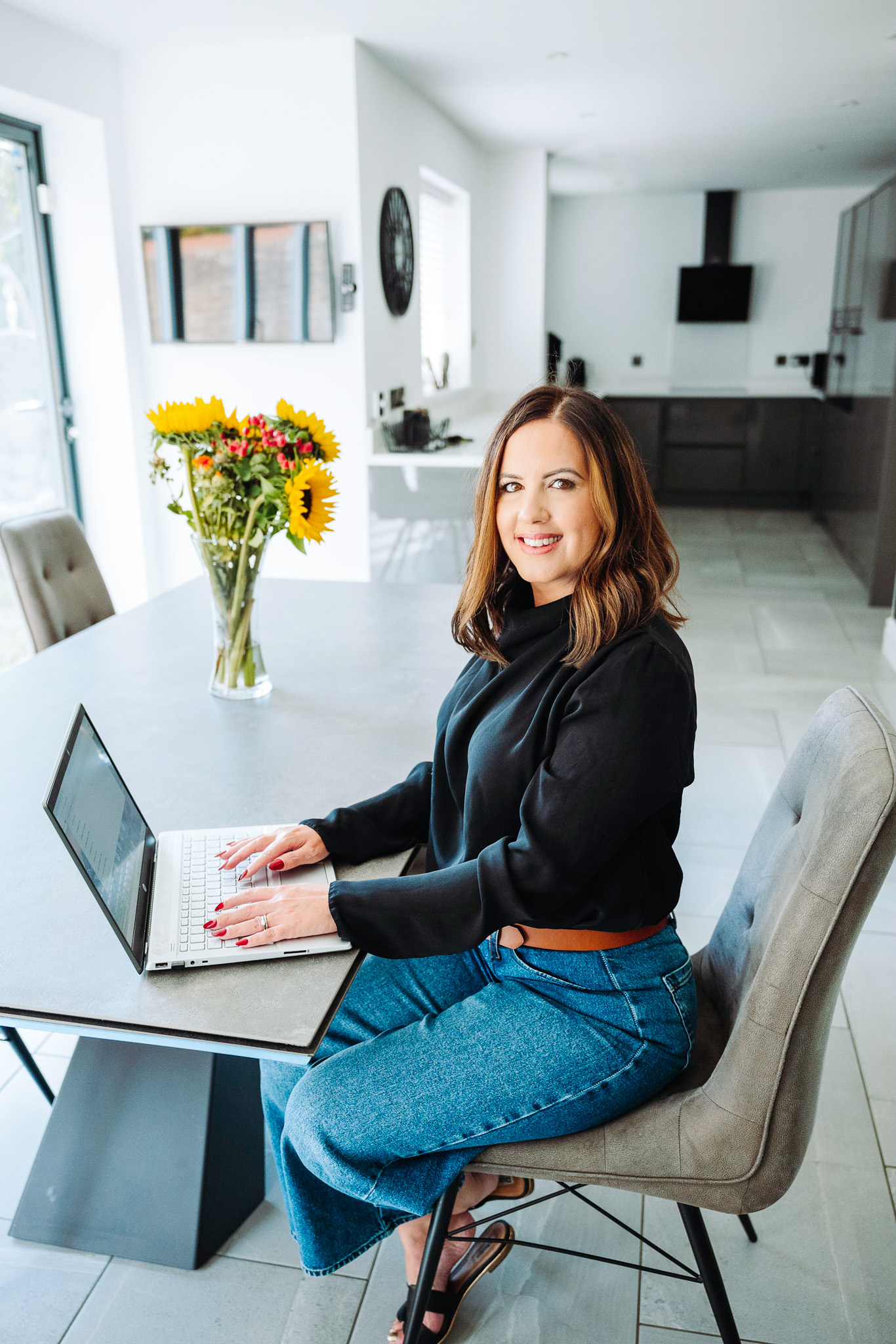 In a bright kitchen, a woman in a black top and jeans sits at a modern dining table using her laptop.