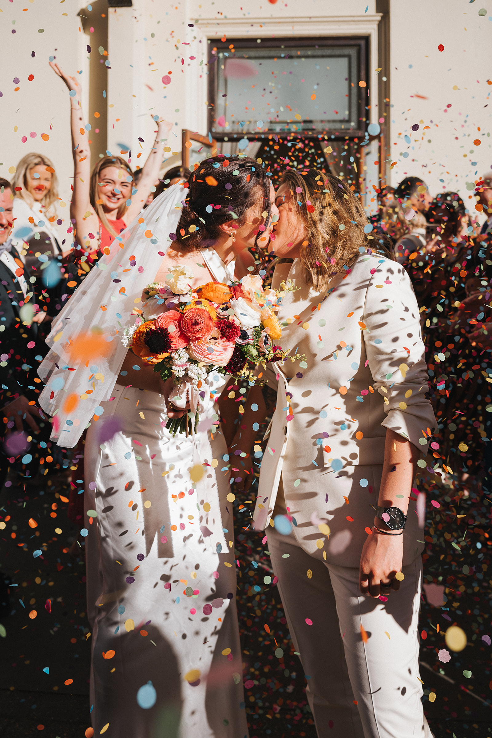 Two brides kissing in colourful confetti outside Brighton Town Hall